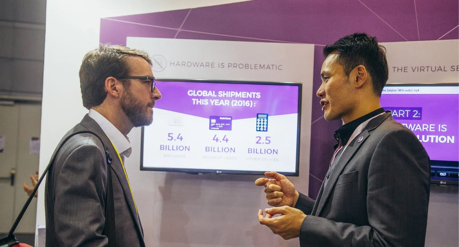 Two men talking at a conference booth in front of a screen of global shipments charts