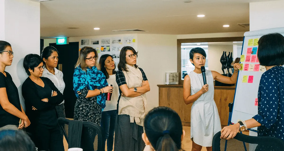 A presenter holding a microphone beside a board of sticky notes while a group watches